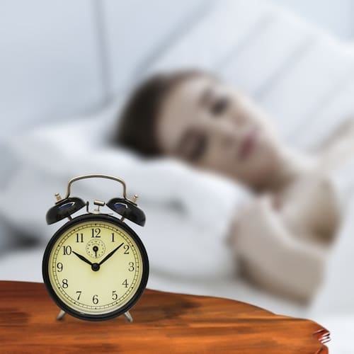 Insomnia Reviews An alarm clock showing 10am and in the background a lady fast asleep in bed